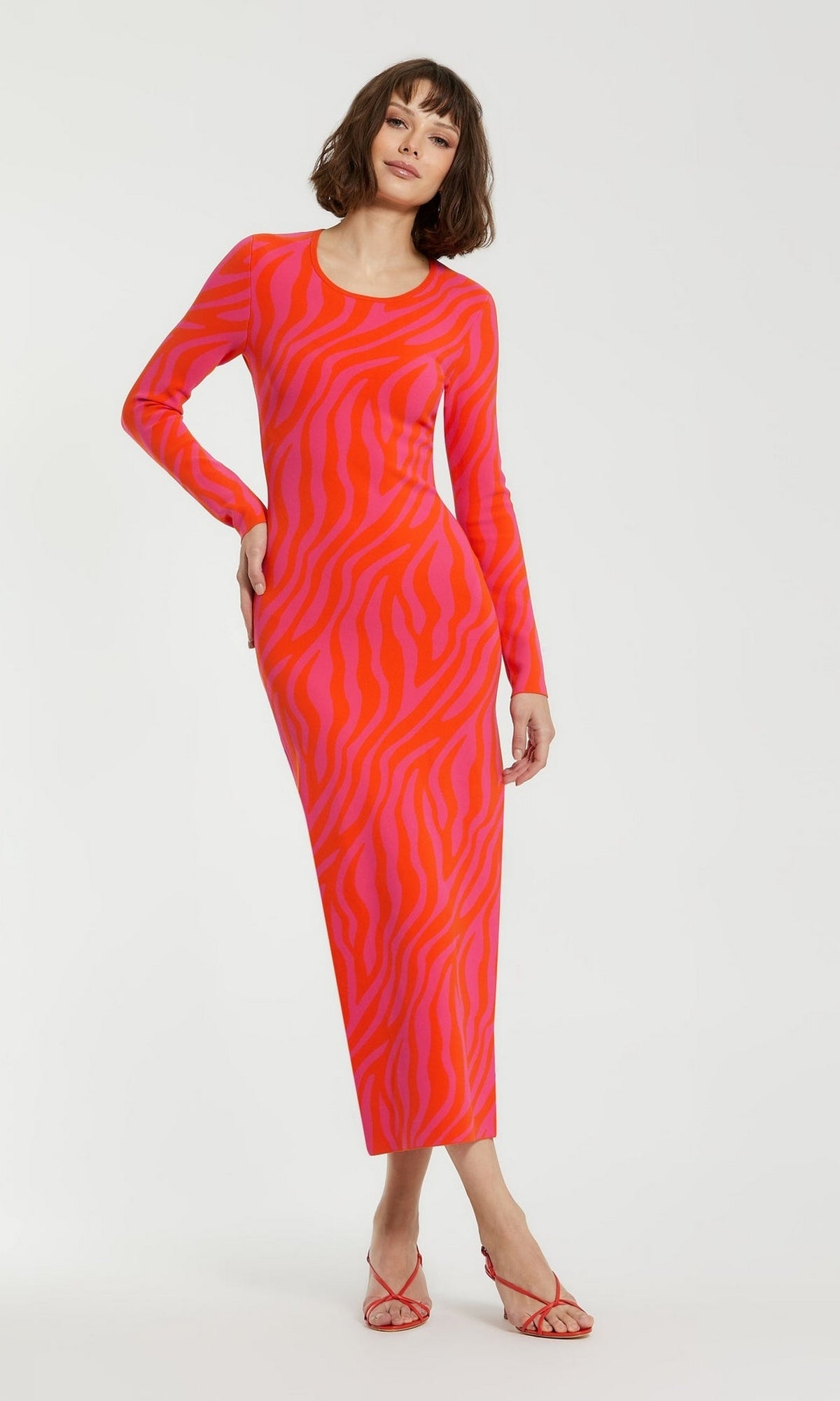 A woman models the Mac Duggal Pink Casual Wedding-Guest Dress K12008, a long-sleeve, ankle-length zebra-print dress with bold red accents and red strappy heels, posed against a white background for a standout occasion look.